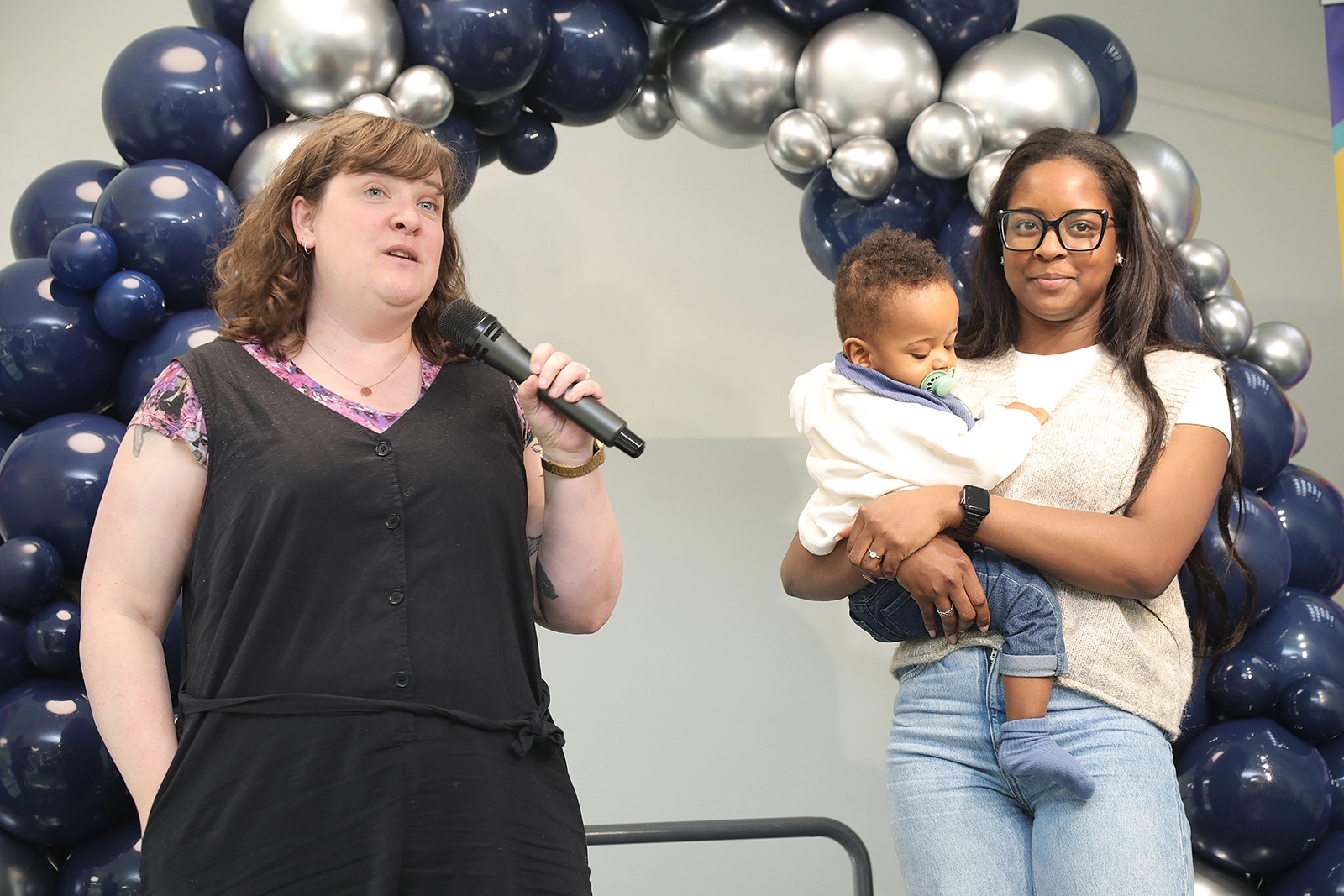 A woman with light skin and brown hair speaks into a microphone next to another woman with dark skin and long hair holding a baby with a pacifier, standing in front of a decorative arch of navy blue and silver balloons.