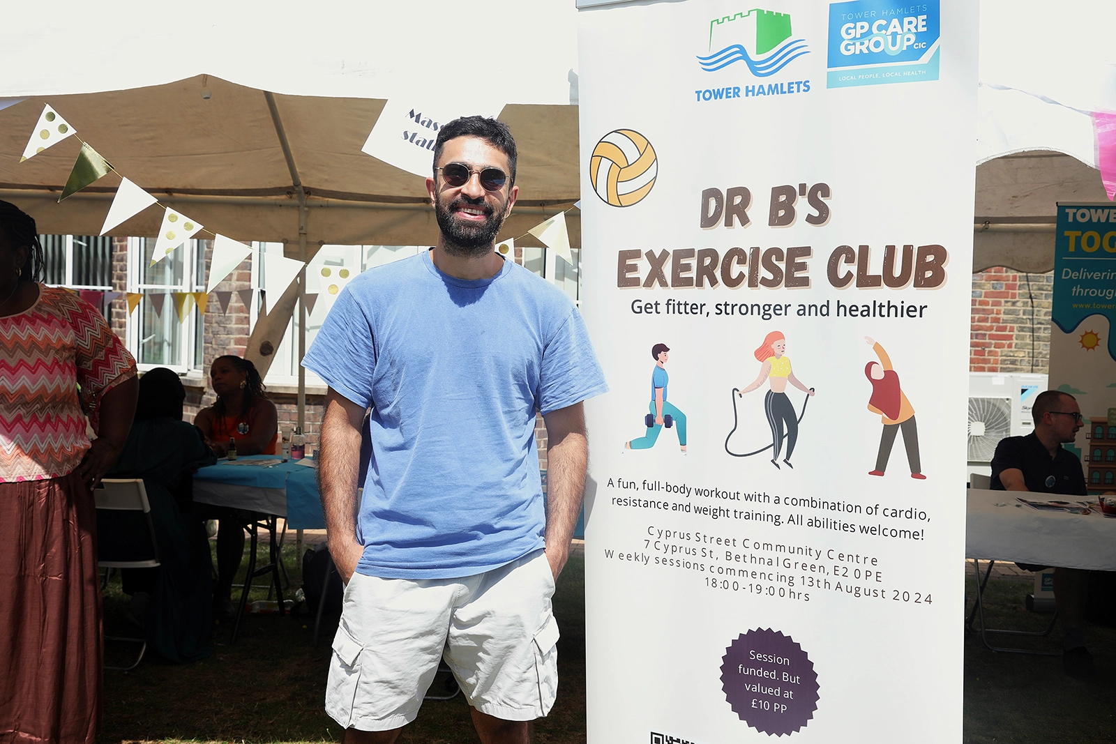 Man wearing sunglasses, a blue t-shirt, and white shorts standing smiling outdoors next to a banner for Dr B's Exercise Club, promoting a fun full-body workout with cardio, resistance, and weight training at Cyprus Street Community Centre.
