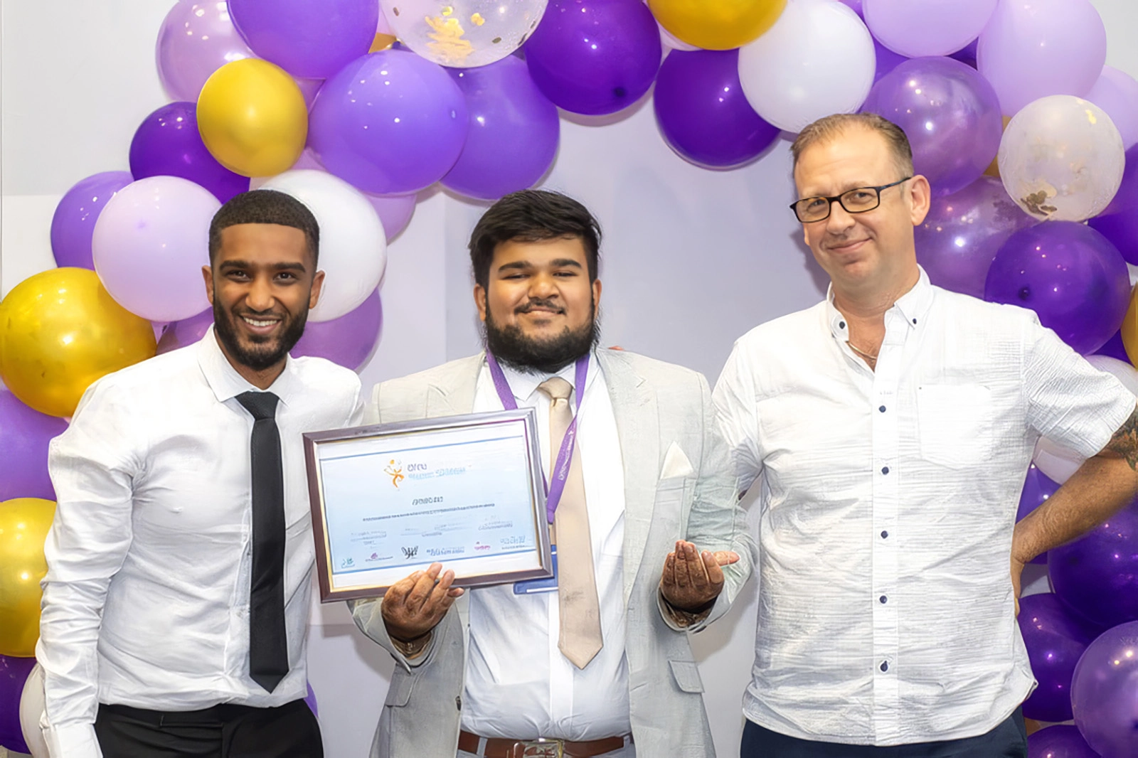 Three men standing in front of a purple and gold balloon arch, the man in the center holding a framed certificate and a glass award, all smiling.