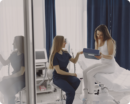 Healthcare professional in navy scrubs showing a clipboard to a woman sitting on an exam table in a medical office.