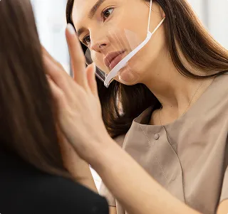 Woman wearing a transparent face shield under her nose, closely applying makeup to another person.
