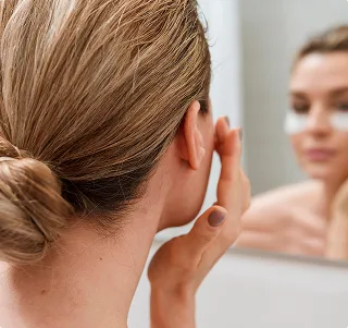 Woman with brown hair tied in a bun applying skincare cream to her face while looking in a mirror.