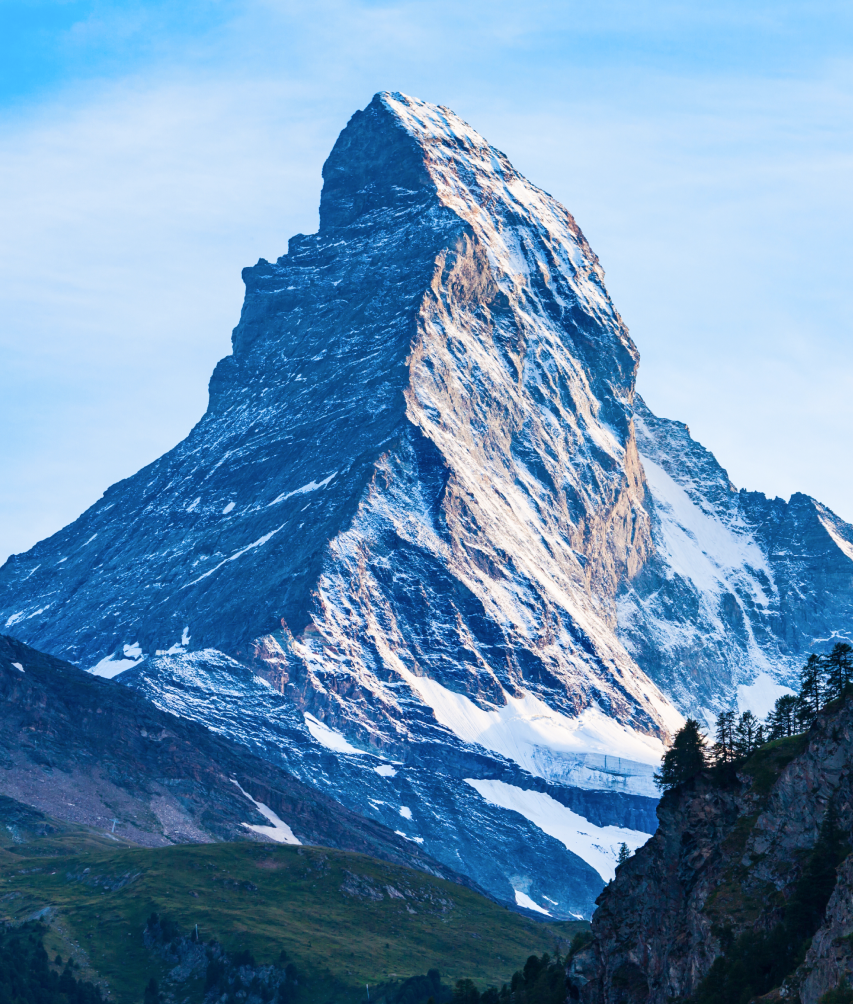 Sunlit peak of the snow-covered Matterhorn mountain with green foothills and clear blue sky.