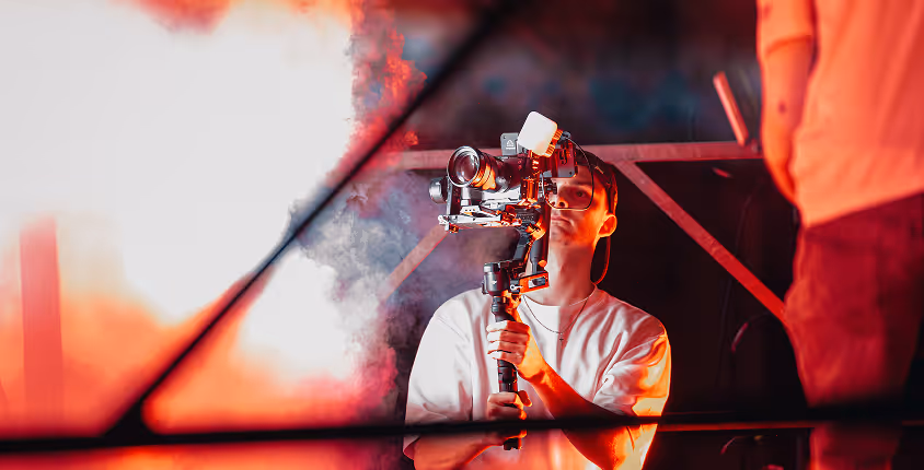 Jeune homme utilisant une caméra stabilisée avec un cadre métallique et une lumière LED dans un environnement embué aux tons rouges.