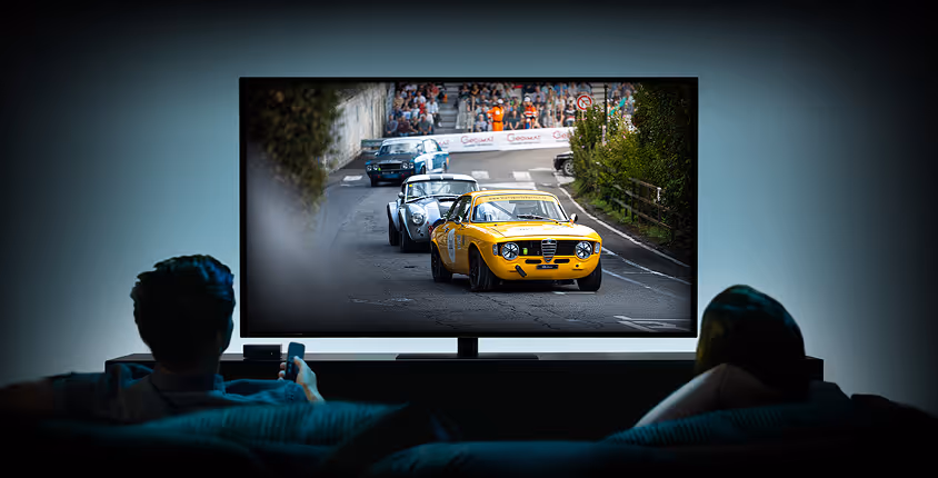 Deux personnes regardant une course de voitures classiques projetée sur un grand écran télévisé dans une pièce sombre.