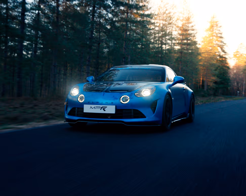 Voiture de sport Alpine bleue avec phares allumés roulant sur une route bordée d'arbres au coucher du soleil.