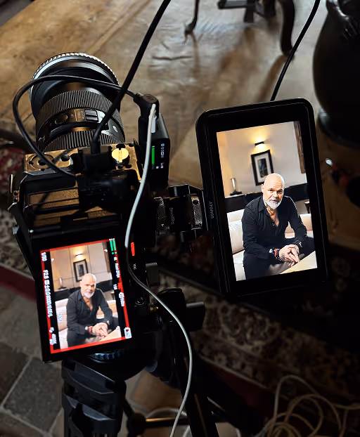 Vue rapprochée d'une caméra professionnelle sur un trépied montrant l'écran avec un homme chauve portant une chemise noire, assis sur un canapé.