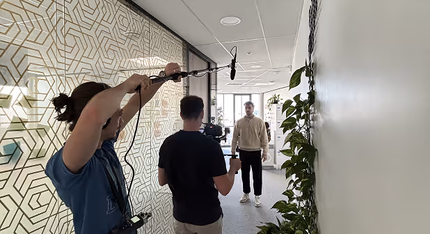 Trois personnes filment une interview dans un couloir, une tient un micro perche, une autre une caméra, et la troisième personne est interviewée.