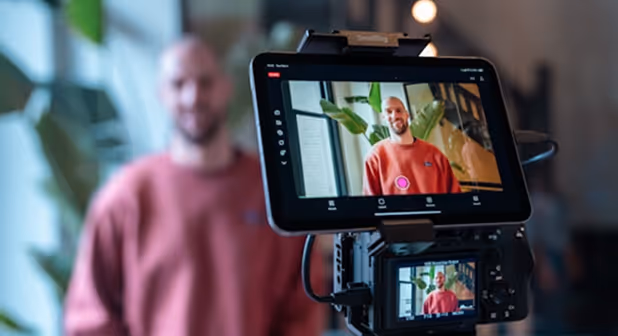 Un homme en sweat-shirt rouge souriant est filmé, avec son image nette visible sur un écran de caméra.