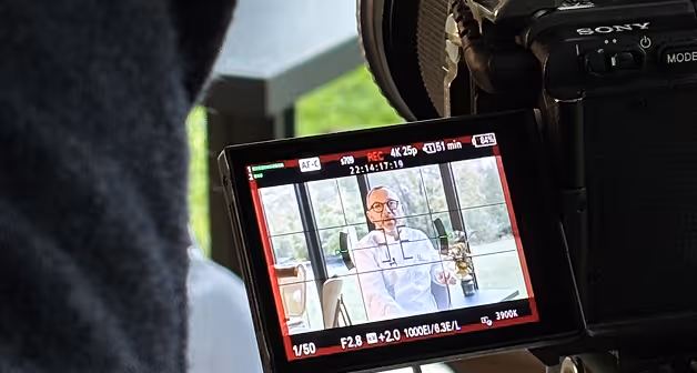 Écran LCD d'un appareil photo Sony montrant un homme en chemise blanche assis devant une grande fenêtre, en train d'être filmé.