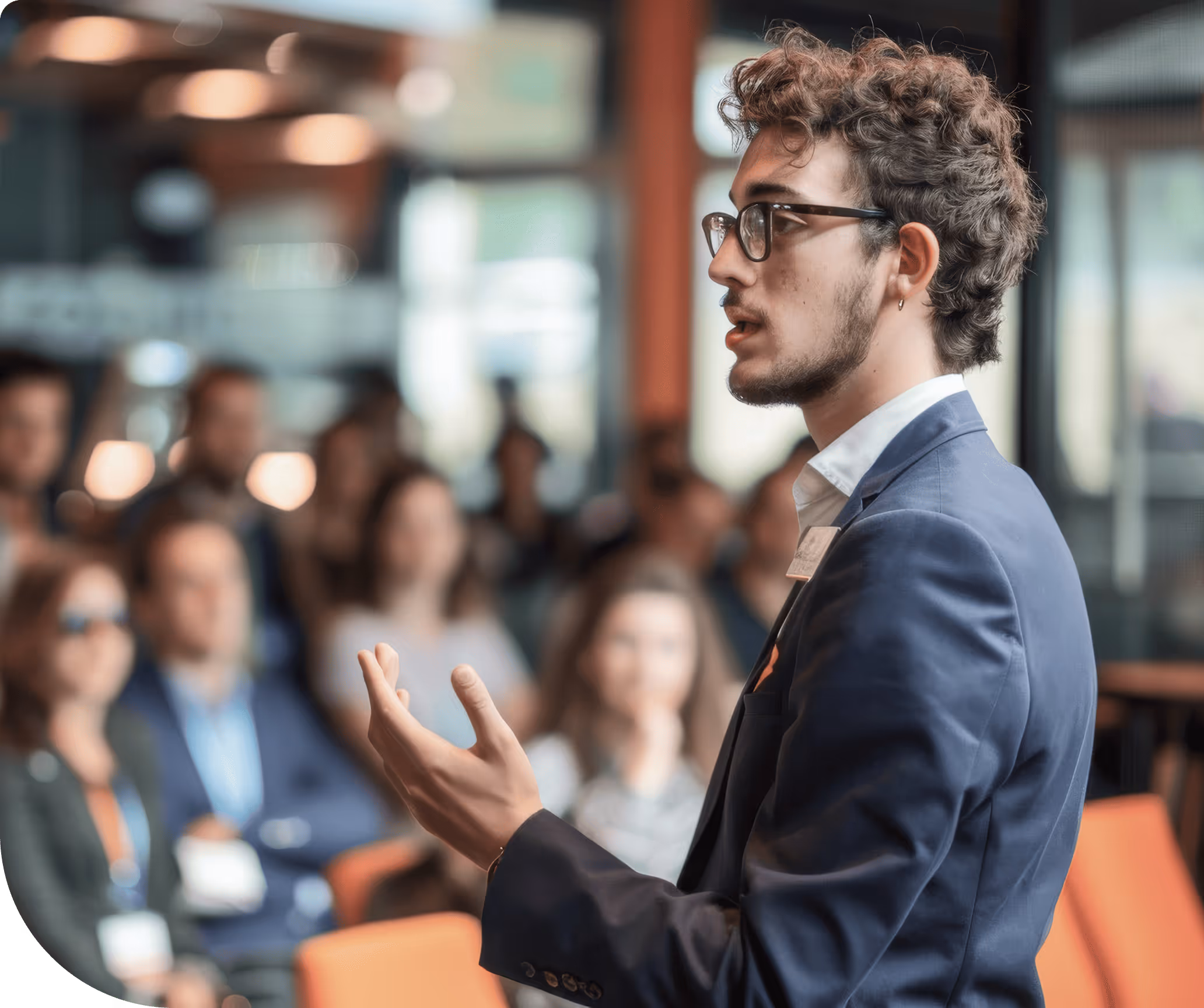 Jeune homme portant des lunettes et un costume bleu parlant devant un public flou lors d'une conférence.