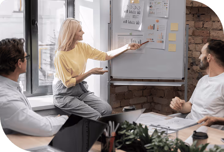 Une femme présente des graphiques sur un tableau blanc à deux collègues assis autour d'une table de réunion.
