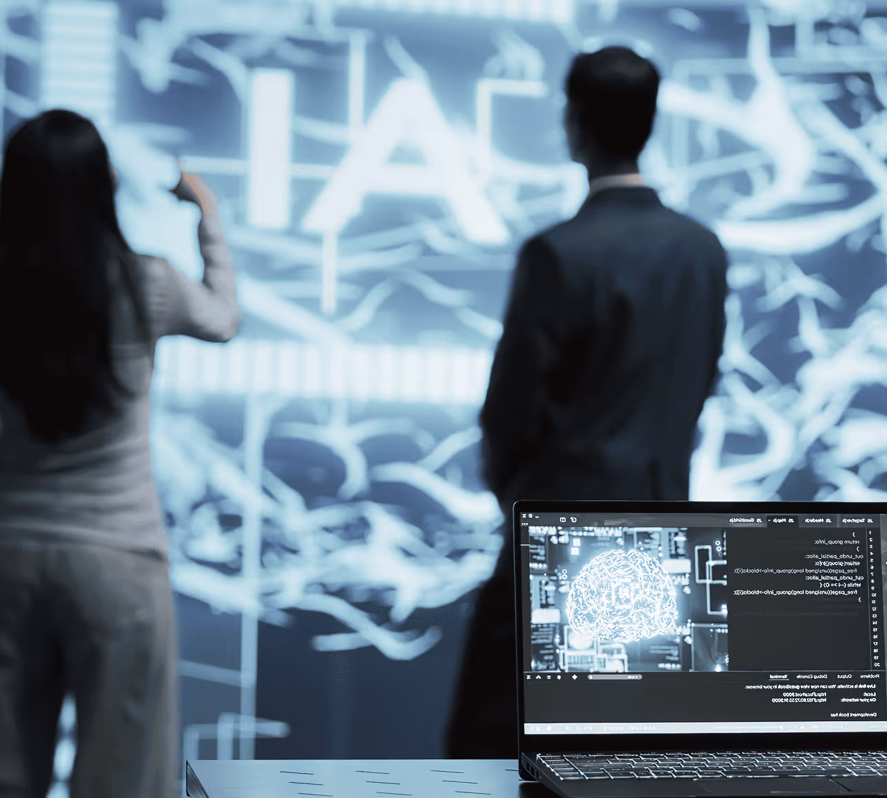 Deux personnes debout face à un grand écran lumineux avec des données abstraites, un ordinateur portable au premier plan affiche une image numérique d'un cerveau.