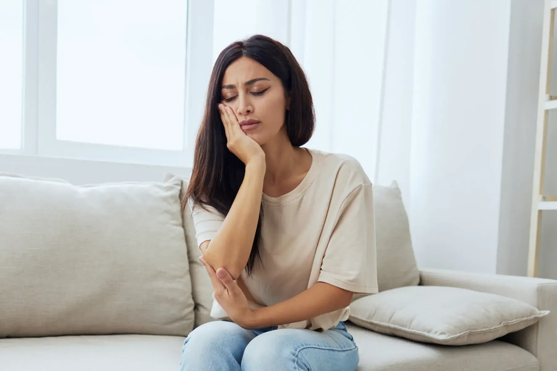 Woman sitting on a sofa holding her cheek and jaw in pain, indicating toothache or discomfort.