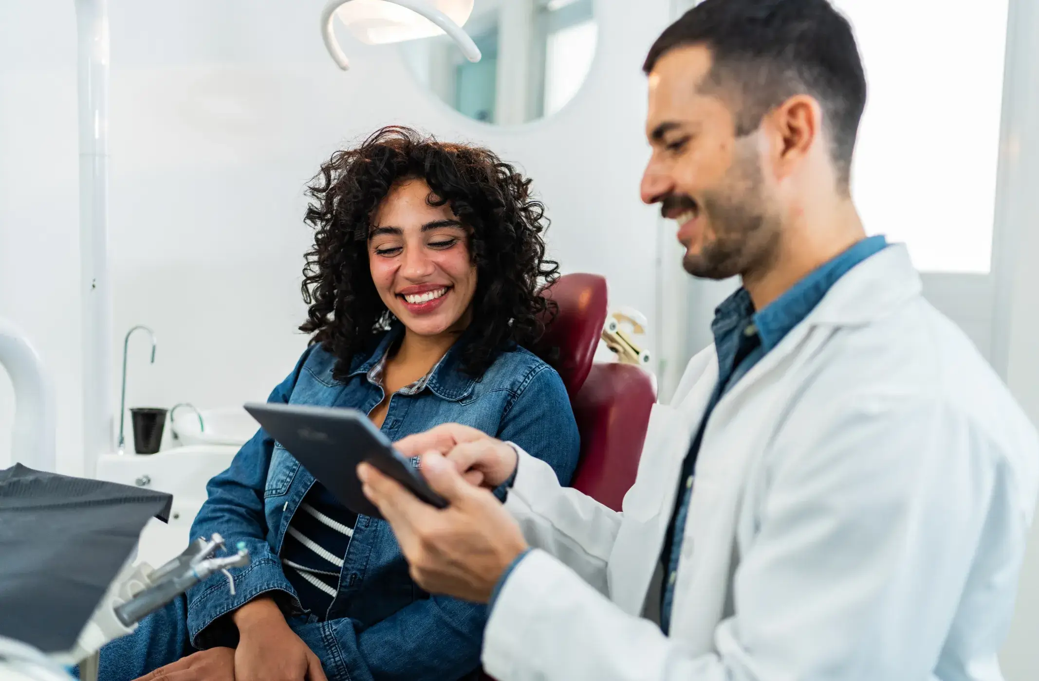 Smiling woman in dental chair looking at a tablet held by a male dentist in a white coat.