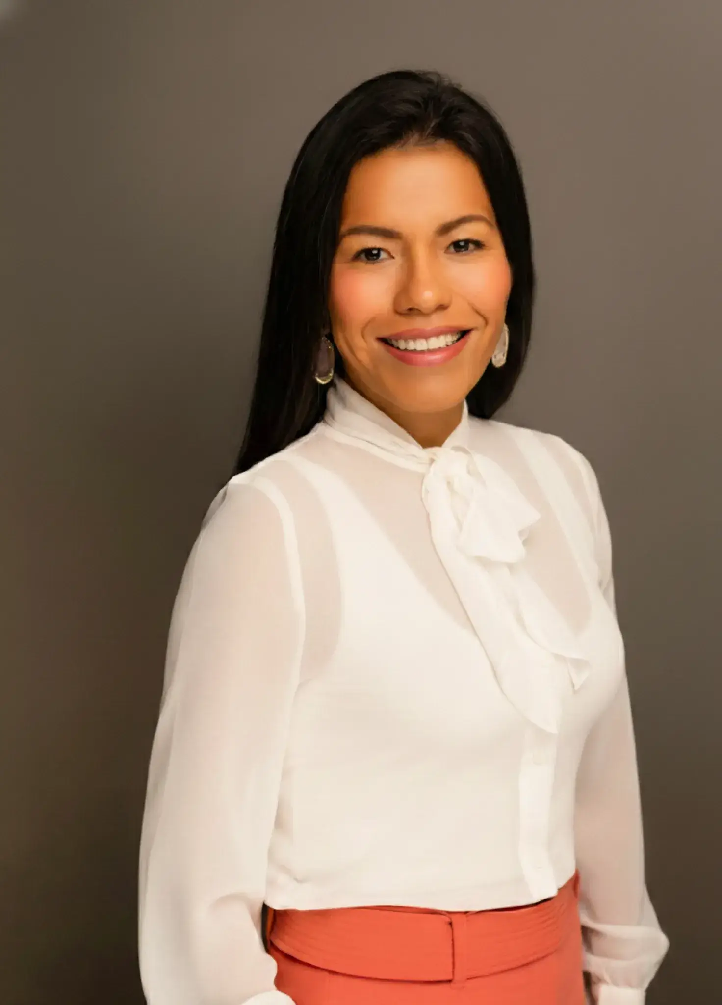 Smiling woman with long dark hair wearing a white blouse with a bow and coral high-waisted pants.