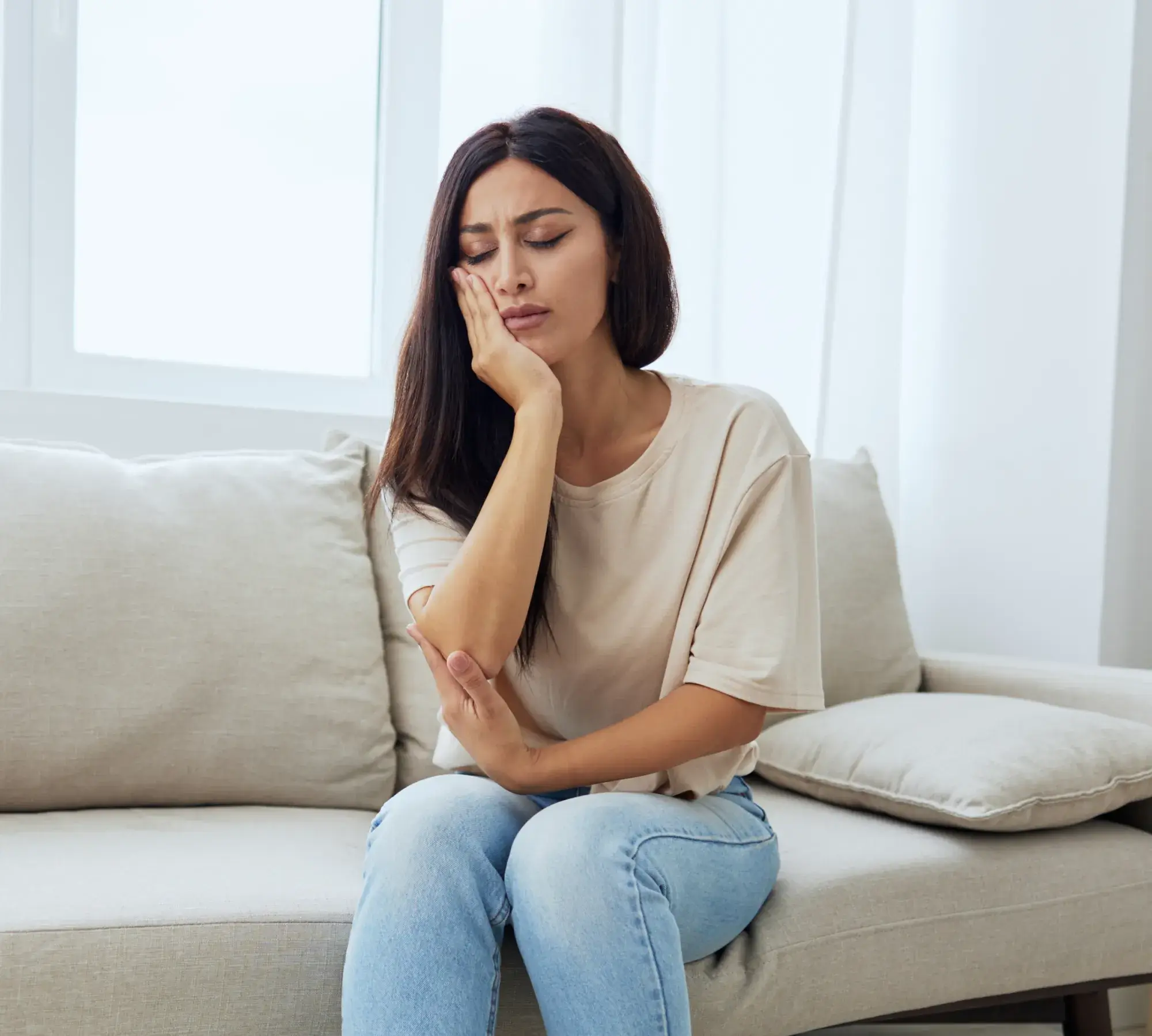 A woman sitting on a couch holding her cheek with a pained expression, indicating toothache or jaw pain.