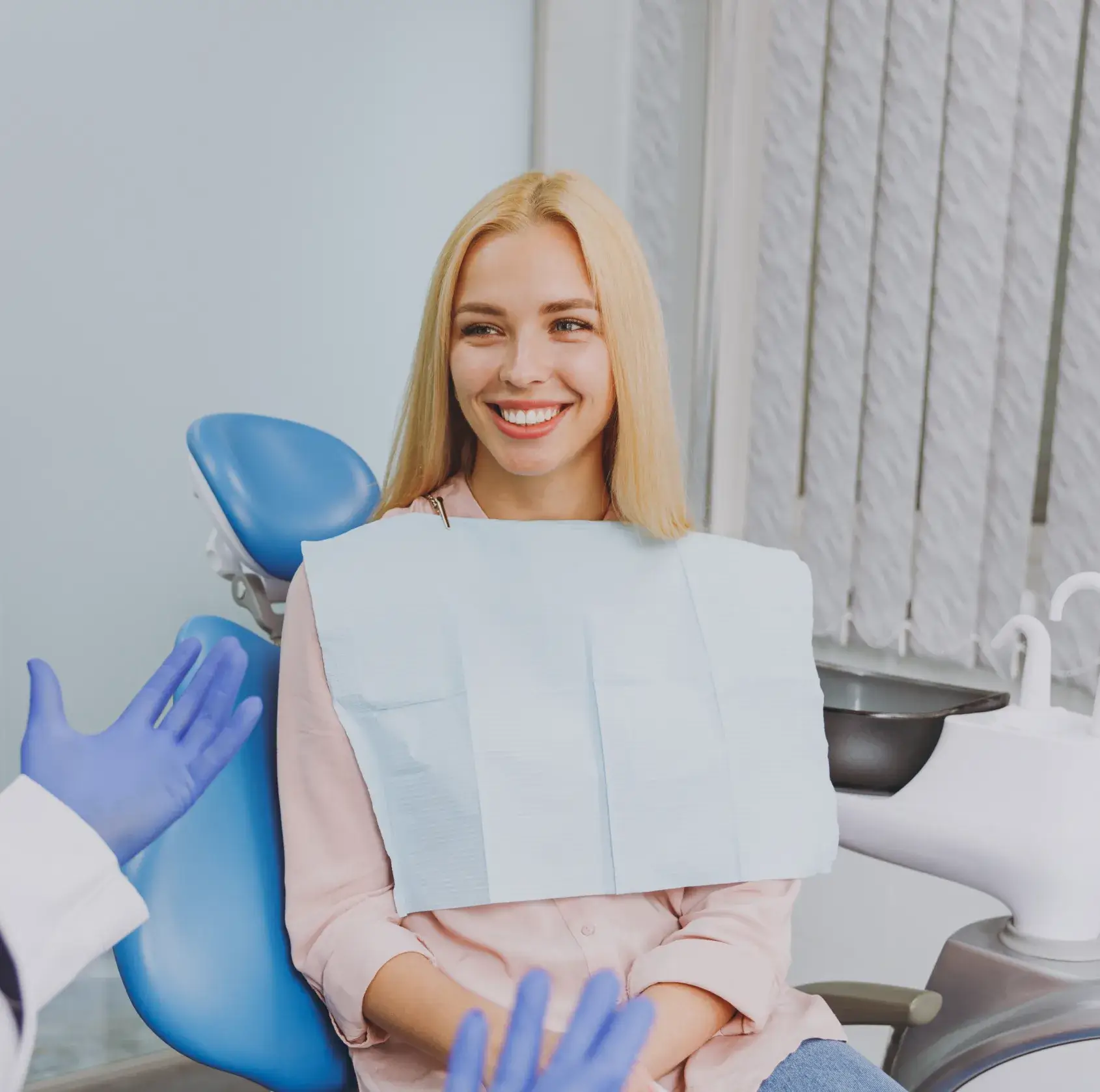 Smiling woman seated in dental chair wearing a protective bib, speaking with a dentist wearing purple gloves.