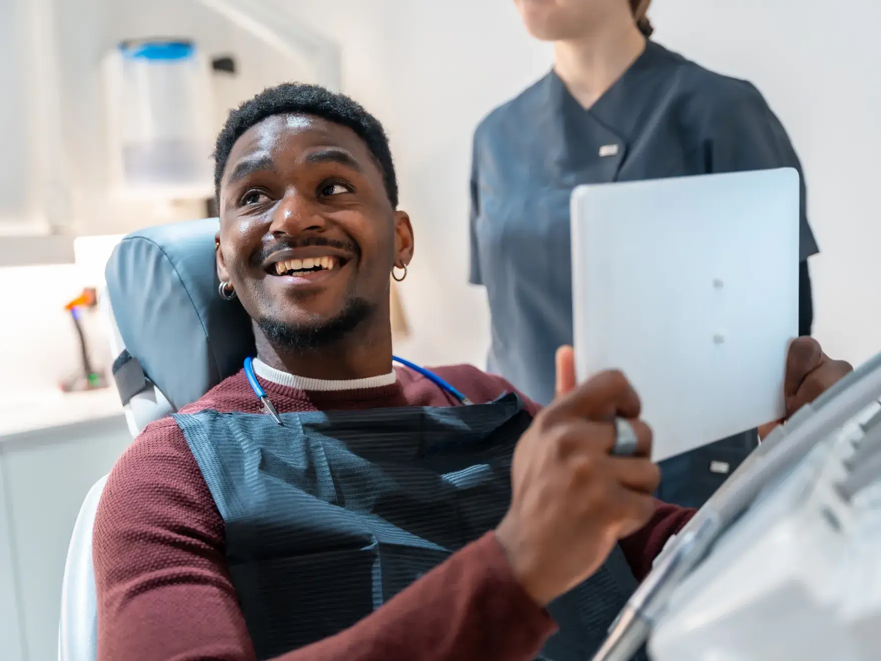 Smiling man sitting in a dental chair holding a mirror with a dental professional standing nearby.