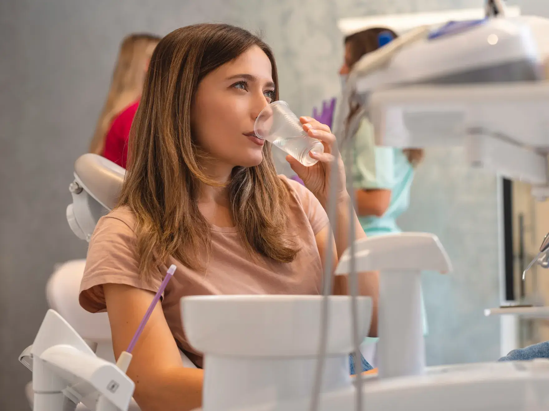 Woman sitting in a dental chair drinking water from a plastic cup, with dental equipment visible in the foreground.