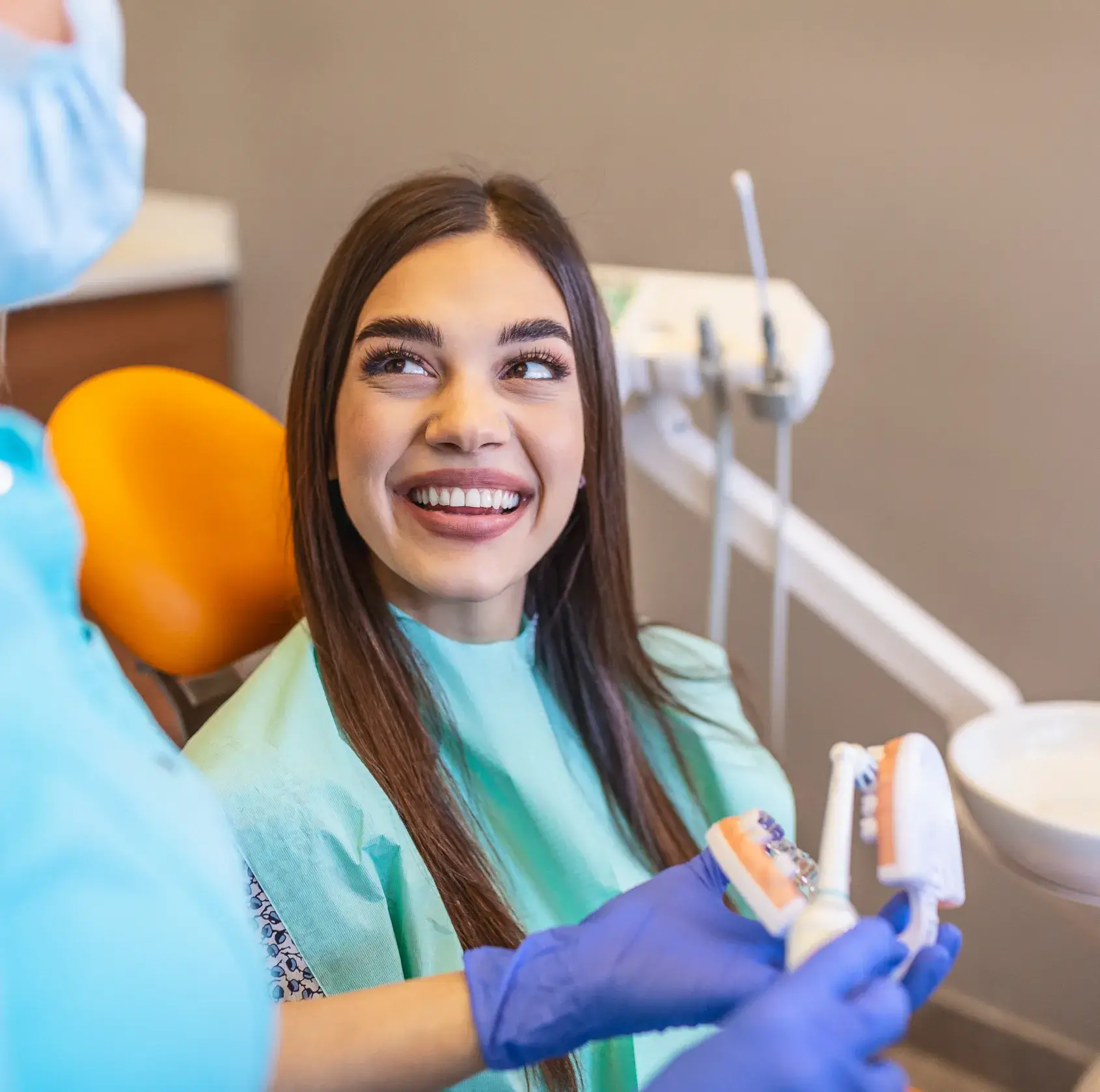 Smiling woman in dental chair looking at dentist wearing blue gloves holding dental models and a toothbrush.