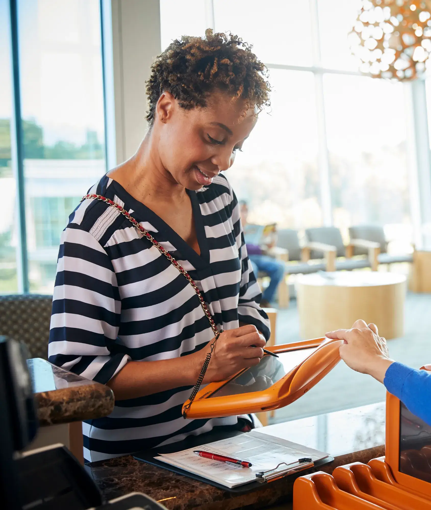 Woman signing on an orange tablet device at a reception desk with a clipboard and pen nearby.
