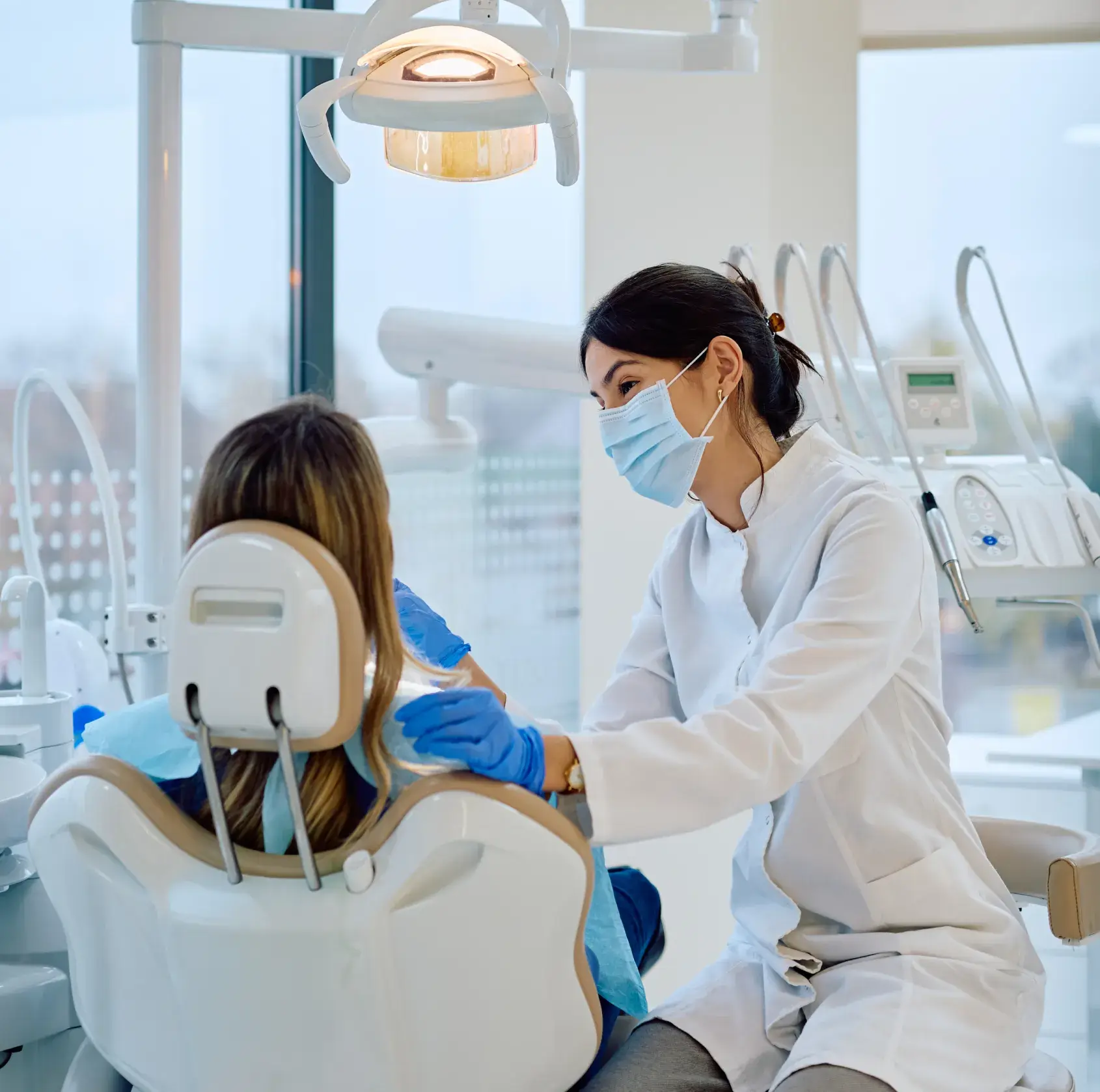 Dentist wearing a mask and gloves smiling and talking to a patient seated in a dental chair in a modern clinic.
