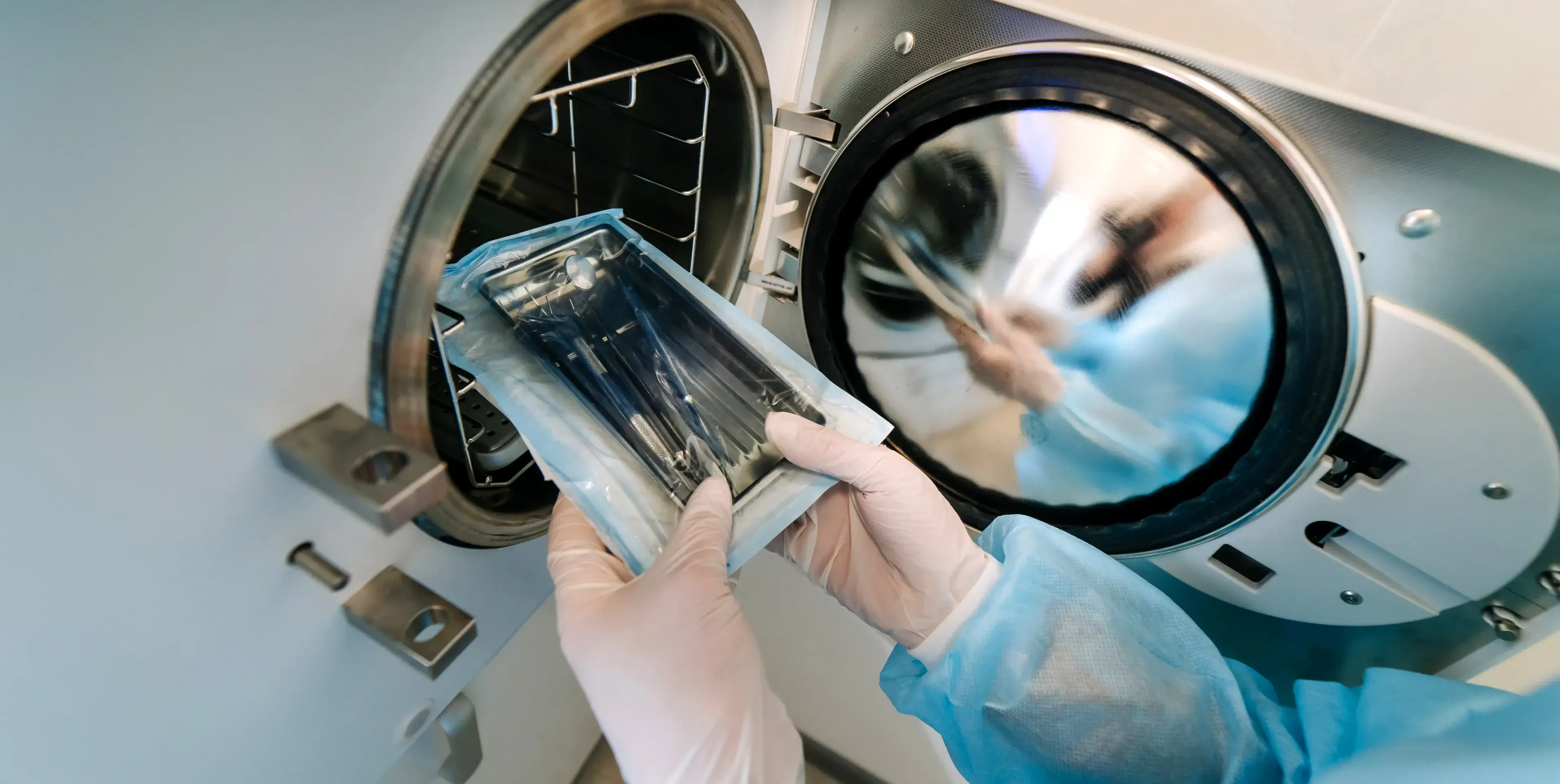 Person in protective gloves and gown placing a sealed tray of sterilized medical instruments into an autoclave sterilizer.