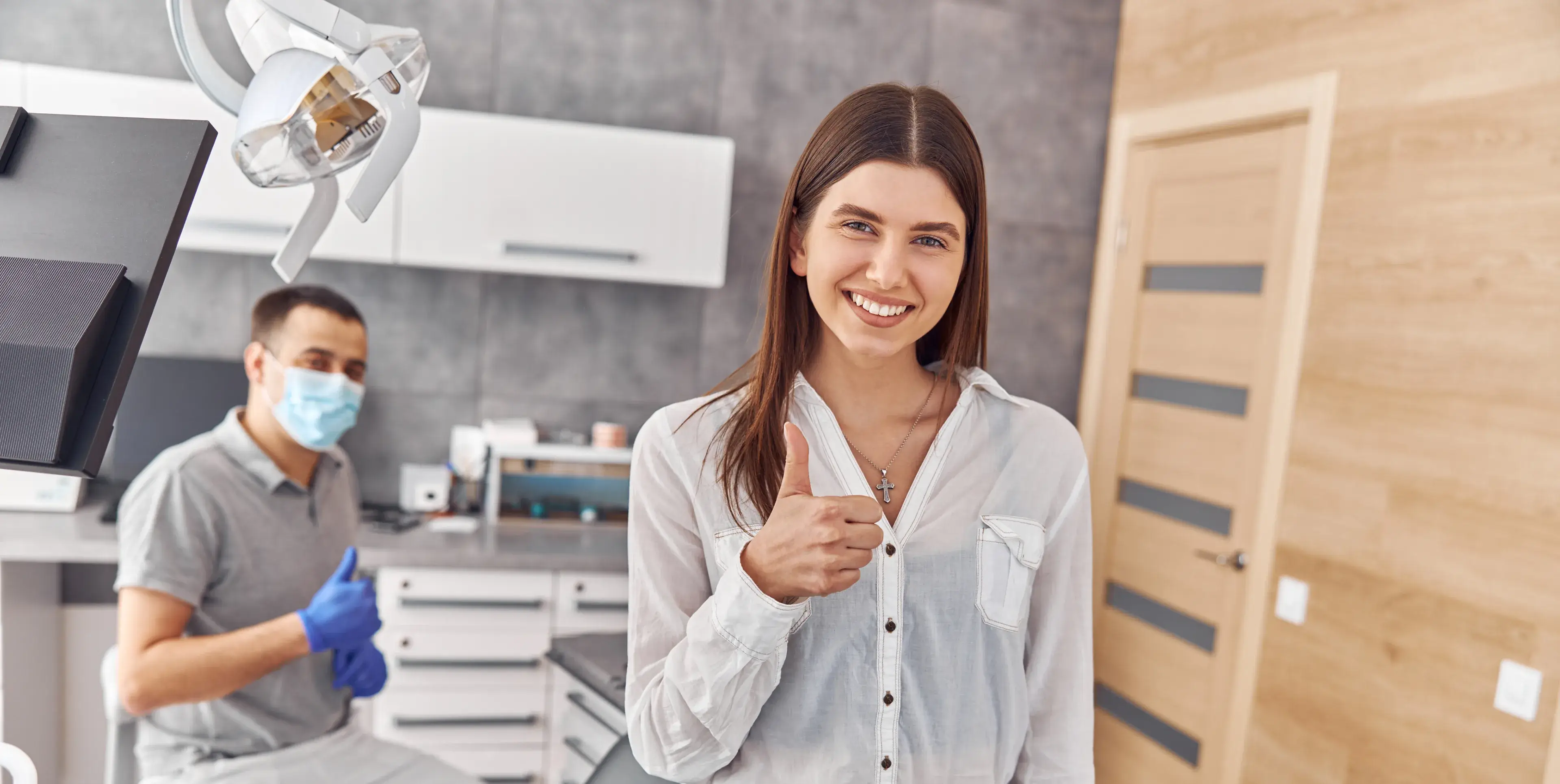Smiling woman giving thumbs up in a dental clinic with a masked dentist also giving a thumbs up in the background.