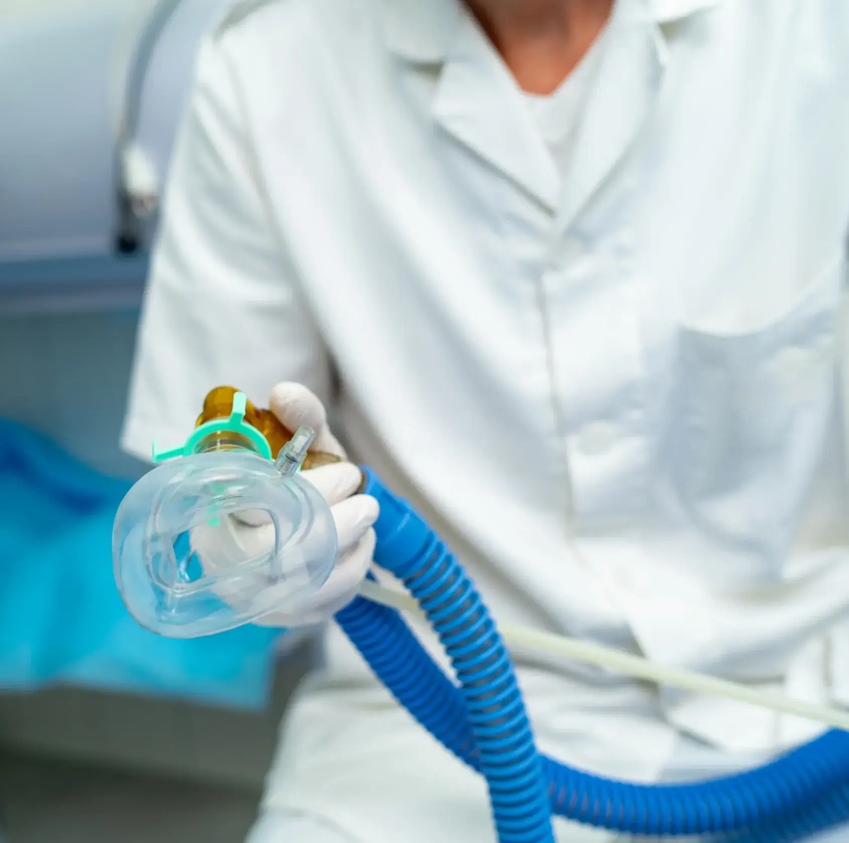 Medical professional in white gloves holding a clear oxygen mask connected to blue tubing.