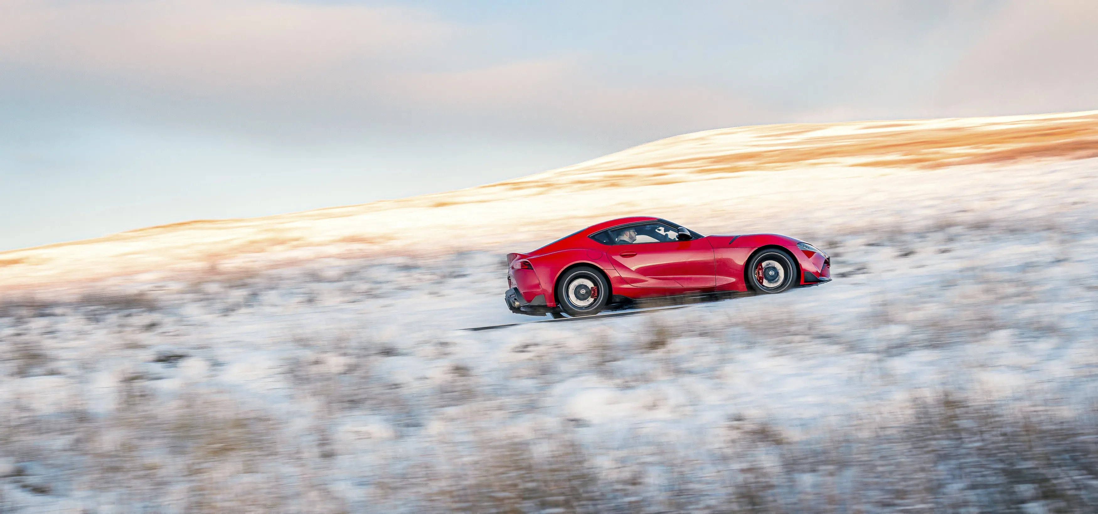 Toyota Supra manual review Rush Magazine - wide angle shot of red A90 Supra driving through Snowdonia National Park framed by snow capped mountains