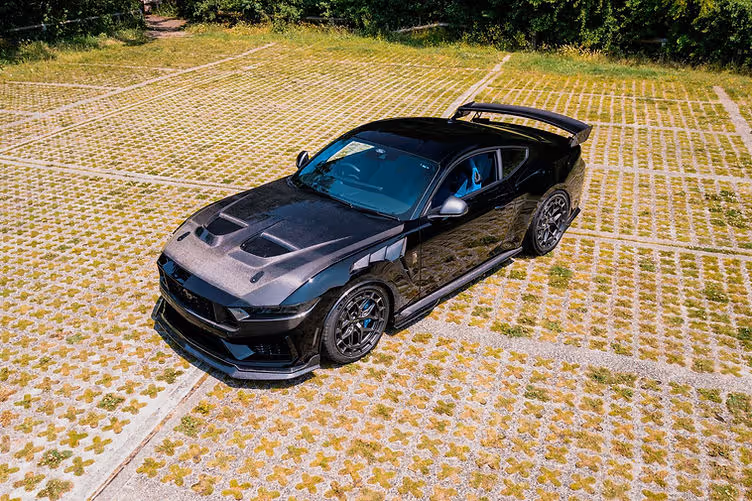 High-angle front three-quarter view of the Clive Sutton CS800DH Mustang, finished in black with carbon fibre bonnet, widebody arches, and large rear wing, parked on a patterned grasscrete surface.