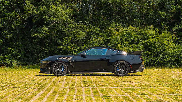 Side profile of the Clive Sutton CS800DH Mustang in black, showcasing its carbon fibre bonnet vents, side skirts, large rear wing, and Vossen wheels, parked on grass with dense green foliage in the background.