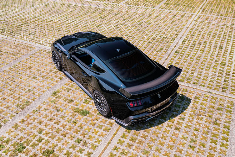 High-angle rear three-quarter view of the Clive Sutton CS800DH Mustang, finished in black with carbon fibre bonnet, widebody arches, and large rear wing, parked on a patterned grasscrete surface.