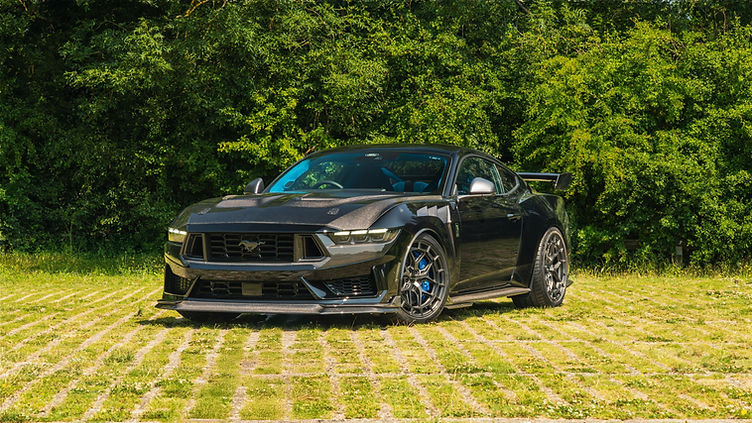 Front view of the Clive Sutton CS800DH Mustang, highlighting its aggressive carbon fibre front splitter, bonnet vents, and widebody stance, parked on a grass-paved surface with dense greenery behind.