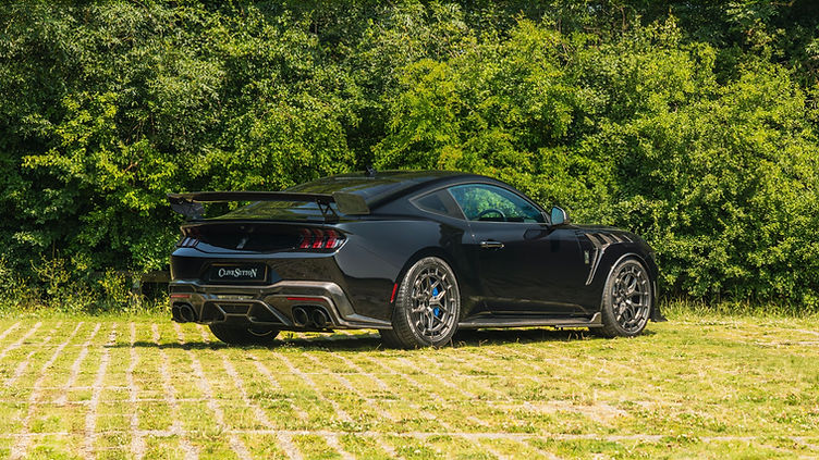 Rear three-quarter view of the Clive Sutton CS800DH Mustang in black, featuring carbon fibre aero parts, quad exhausts, and bronze Vossen wheels, parked on a grass-paved surface with dense greenery in the background.
