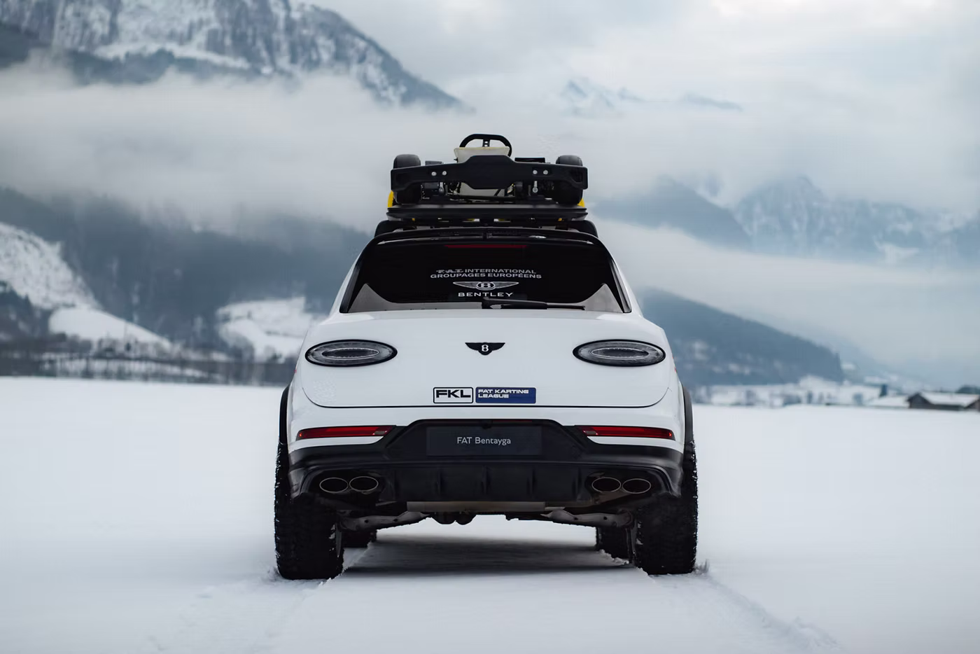 Bentayga X Concept rear static image. White SUV with a go-kart on top in a snowy landscape, mountains in the background. "FAT Bentayga" visible on license plate. Overcast sky.