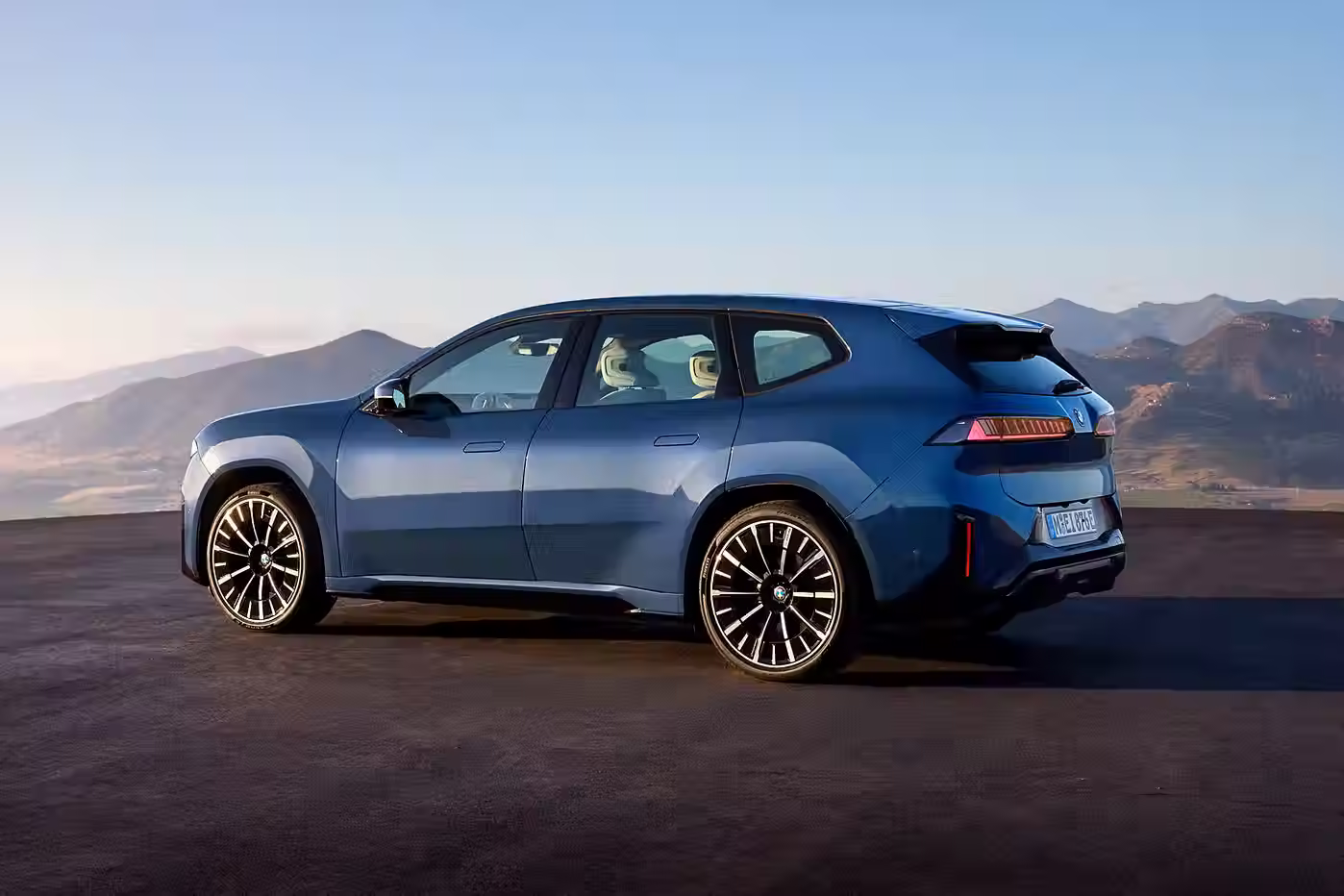 Rear three-quarter view of the BMW iX3 50 xDrive parked on a scenic overlook, showing its sloping roofline, angular LED taillights, and blue exterior.