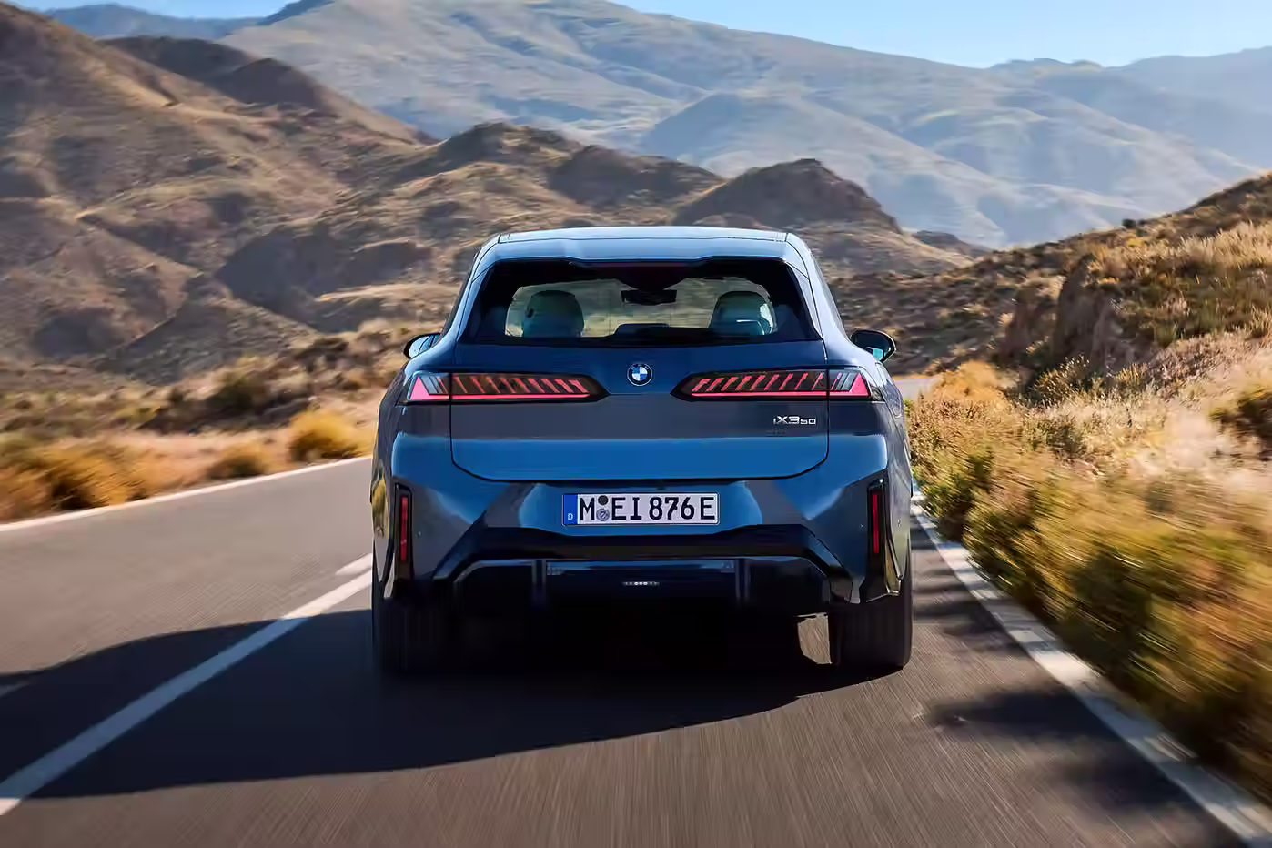 Rear view of a BMW iX3 50 xDrive driving through a winding mountain road, showing its tail-lights, badge, and licence plate with a backdrop of sunlit hills.