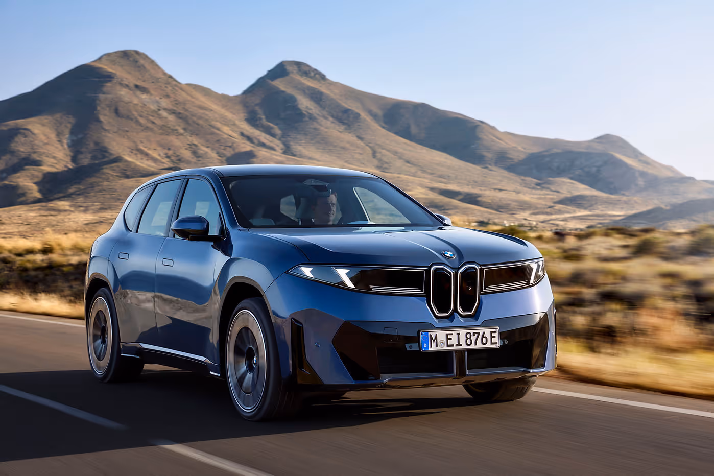 BMW iX3 50 xDrive driving on a mountain road, highlighting its kidney grille, LED headlights, and bold front-end styling.