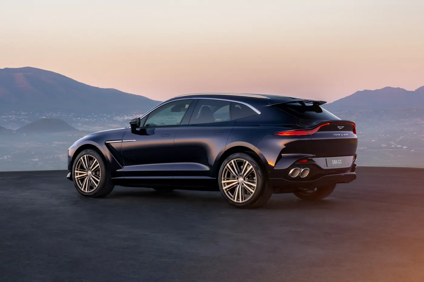 Rear three-quarter view of a dark blue Aston Martin DBX GT parked above a valley at dusk