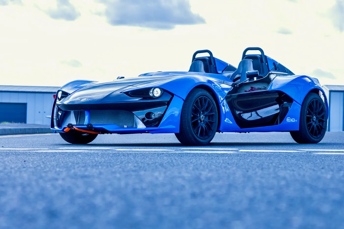 Blue Zenos E10 r2 roadster with black carbon fibre bonnet and black alloy wheels, parked on tarmac, front three-quarter view from low down.