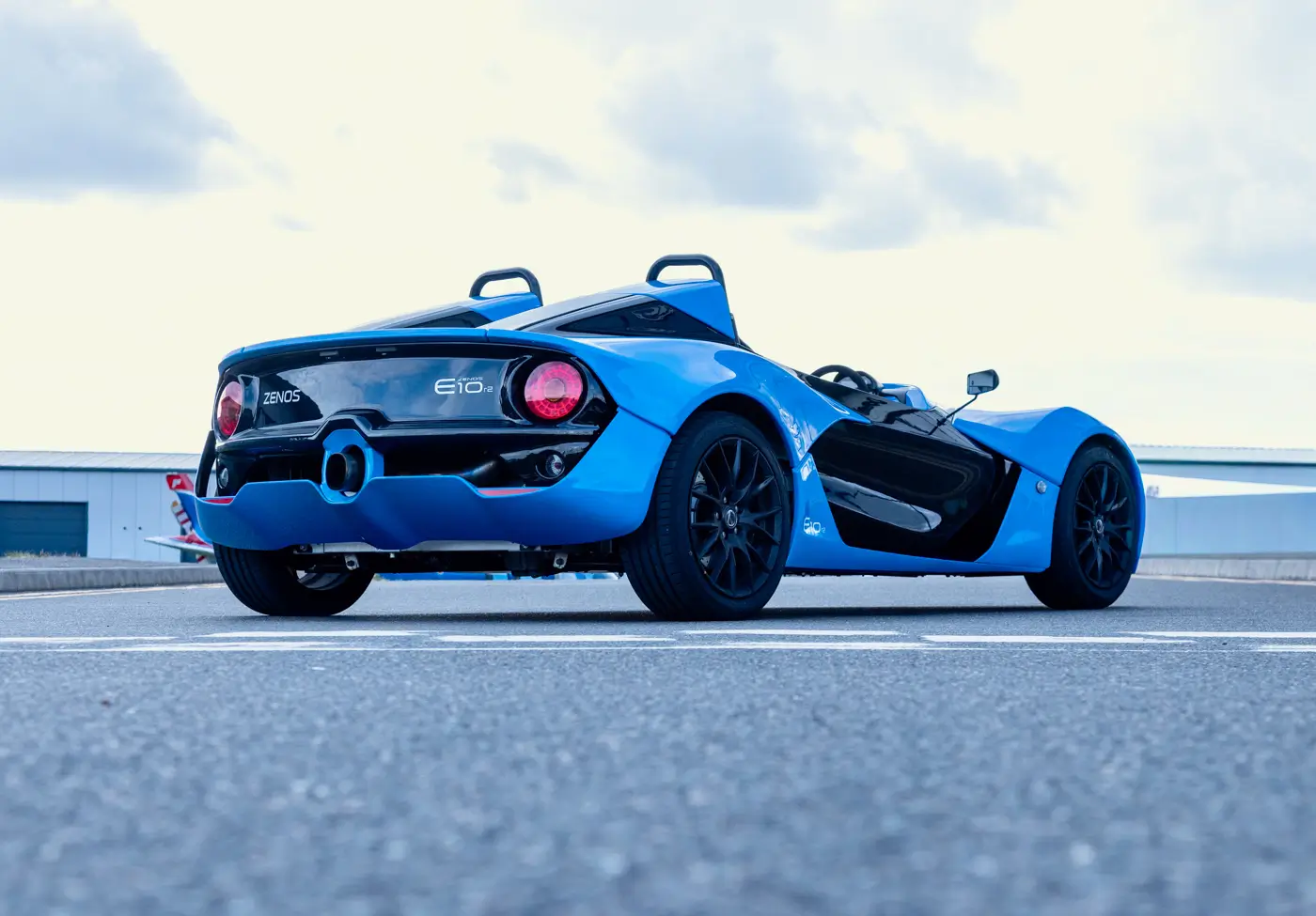 Rear three-quarter view of a blue Zenos E10 r2, showing round tail lights, centre-exit exhaust and twin roll hoops.