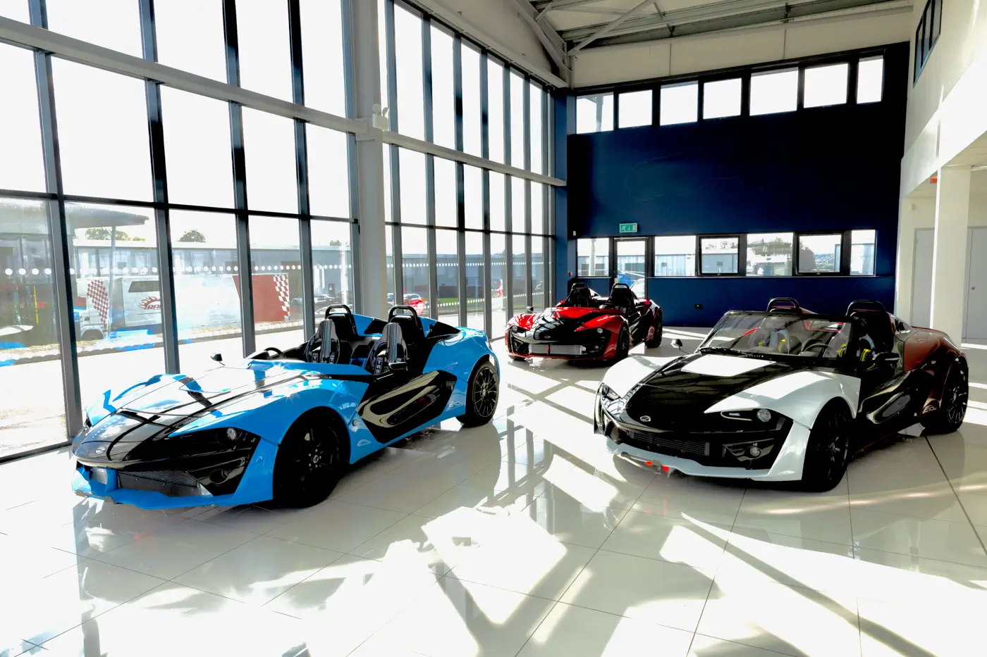 Three Zenos E10 roadsters in blue, red and white, each with black bodywork panels, inside a glass-fronted showroom.