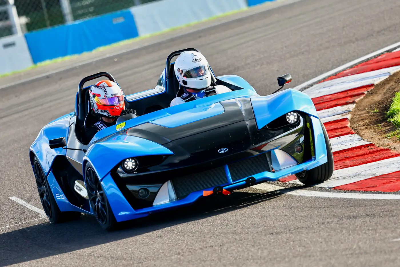 Two helmeted occupants in a blue Zenos E10 cornering on a race circuit beside red and white kerbs.