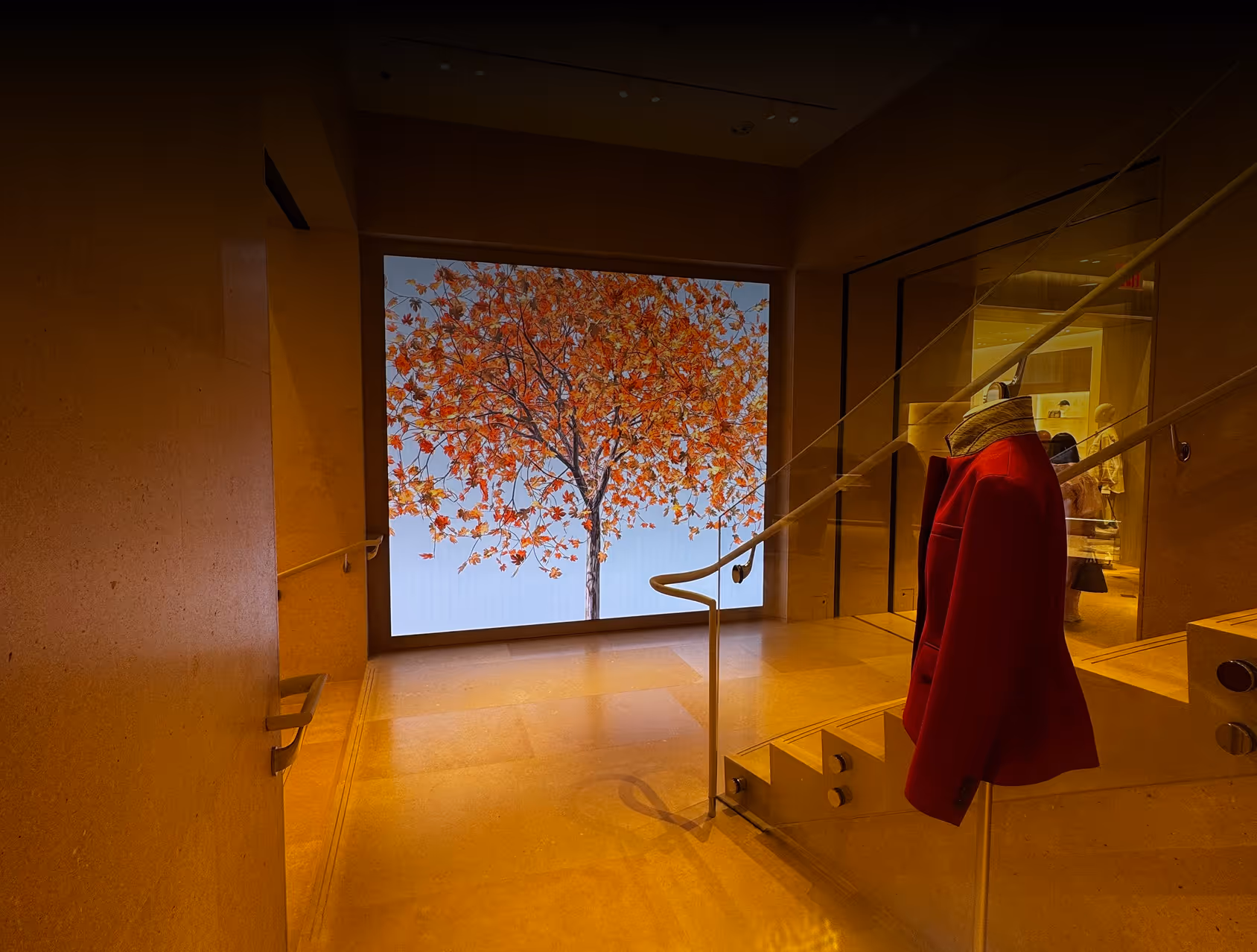 Interior view of a modern store with a large illuminated display of an autumn tree with orange leaves and a red jacket hanging on a glass railing next to stairs.