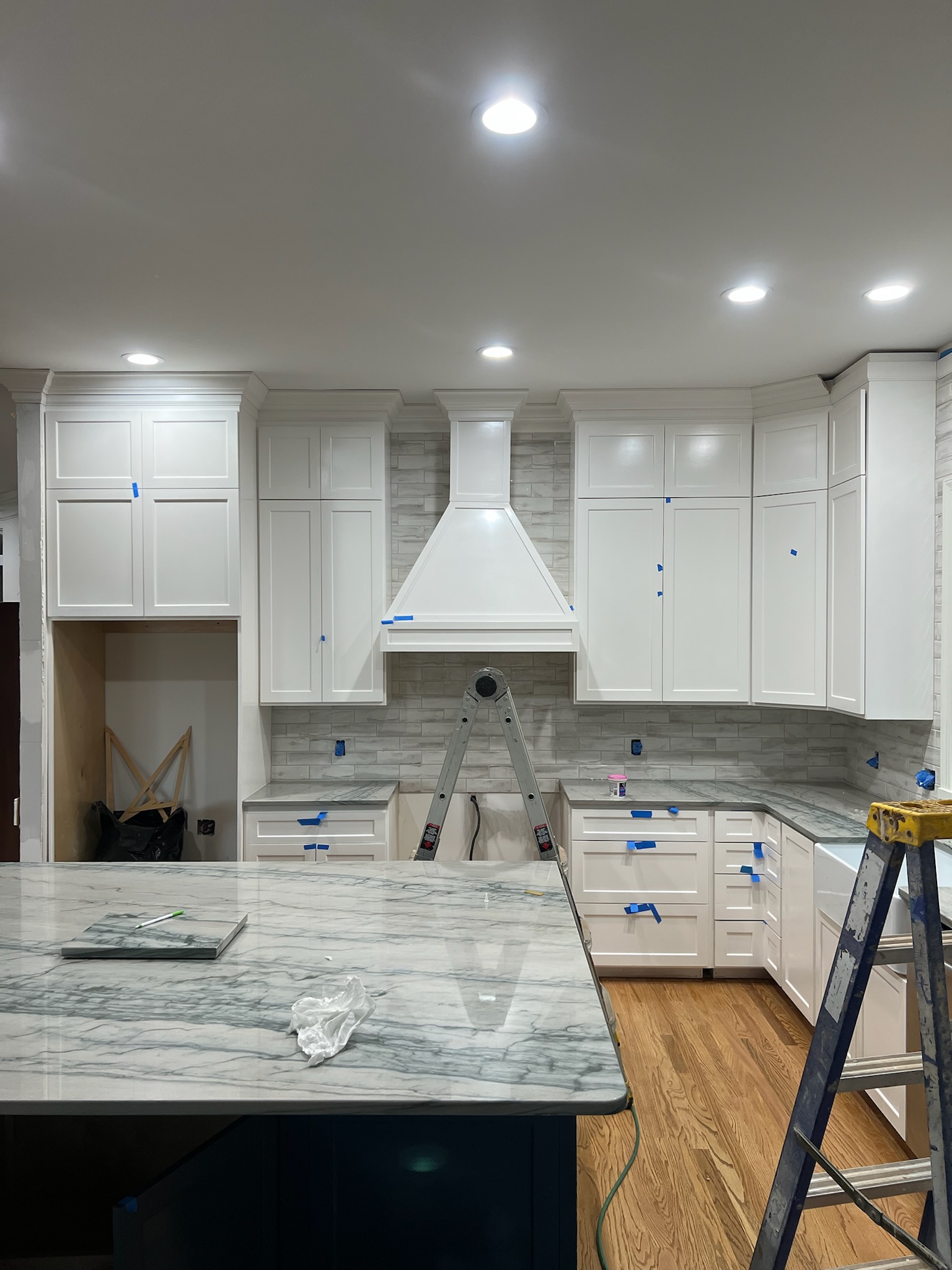 Modern kitchen under construction with white cabinets, marble countertops, gray tiled backsplash, wooden floor, and two ladders.