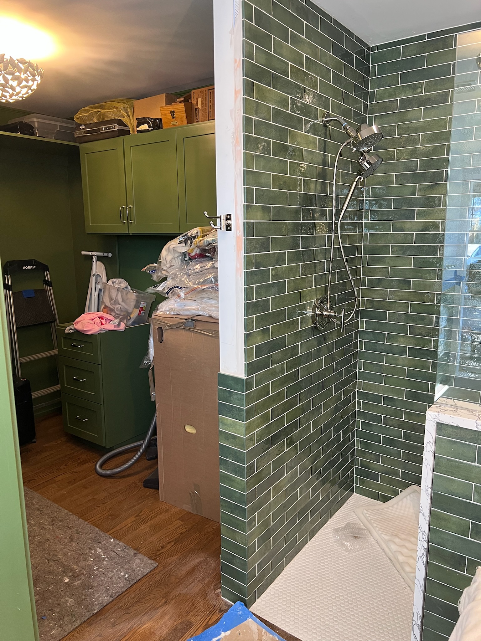 Green-tiled corner shower with chrome showerhead adjacent to a green cabinet storage area with boxes and clothes.