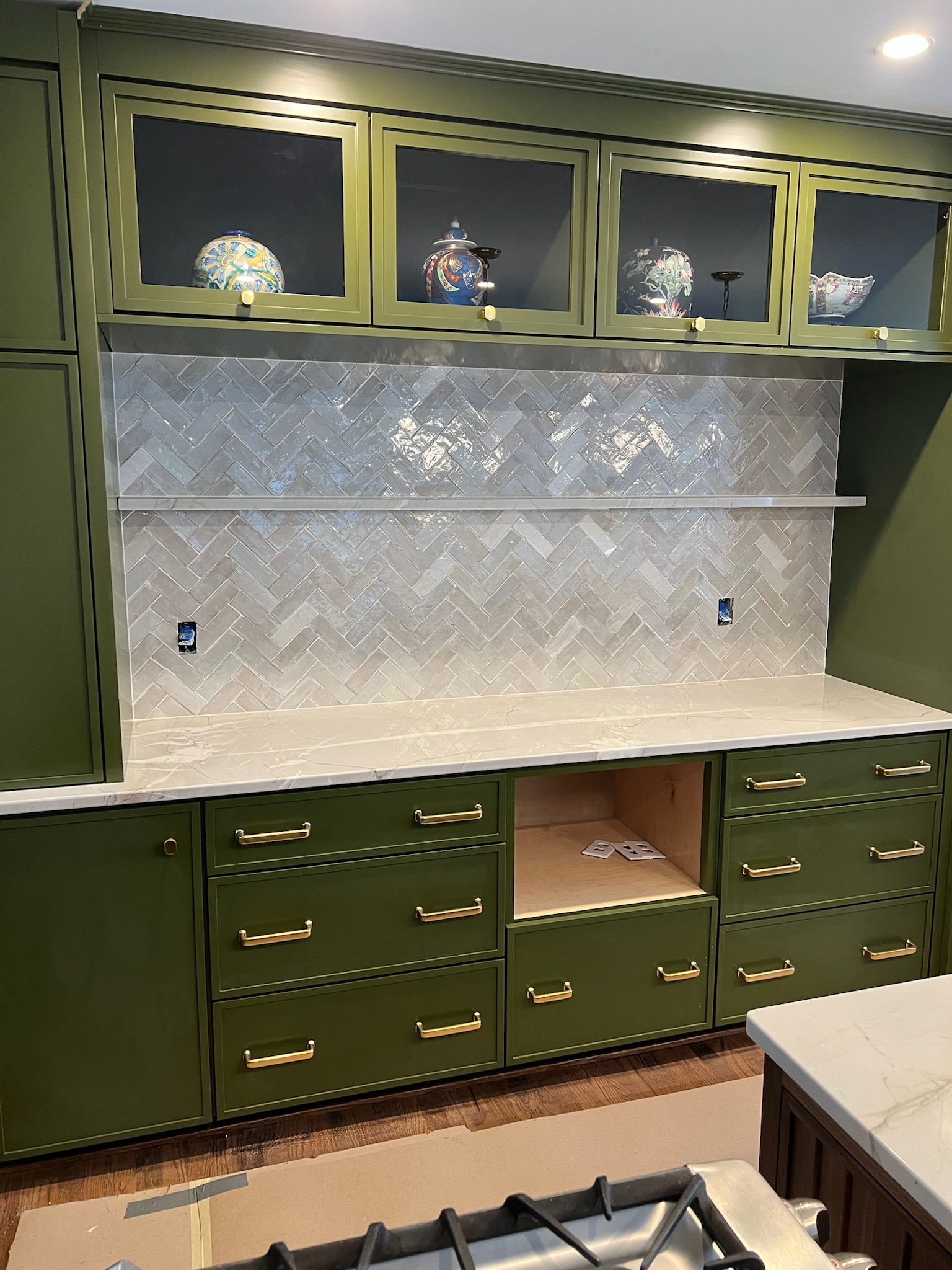 Kitchen cabinetry with green cabinets, brass handles, white marble countertop, herringbone tile backsplash, and upper cabinets with glass doors displaying decorative ceramic vases.