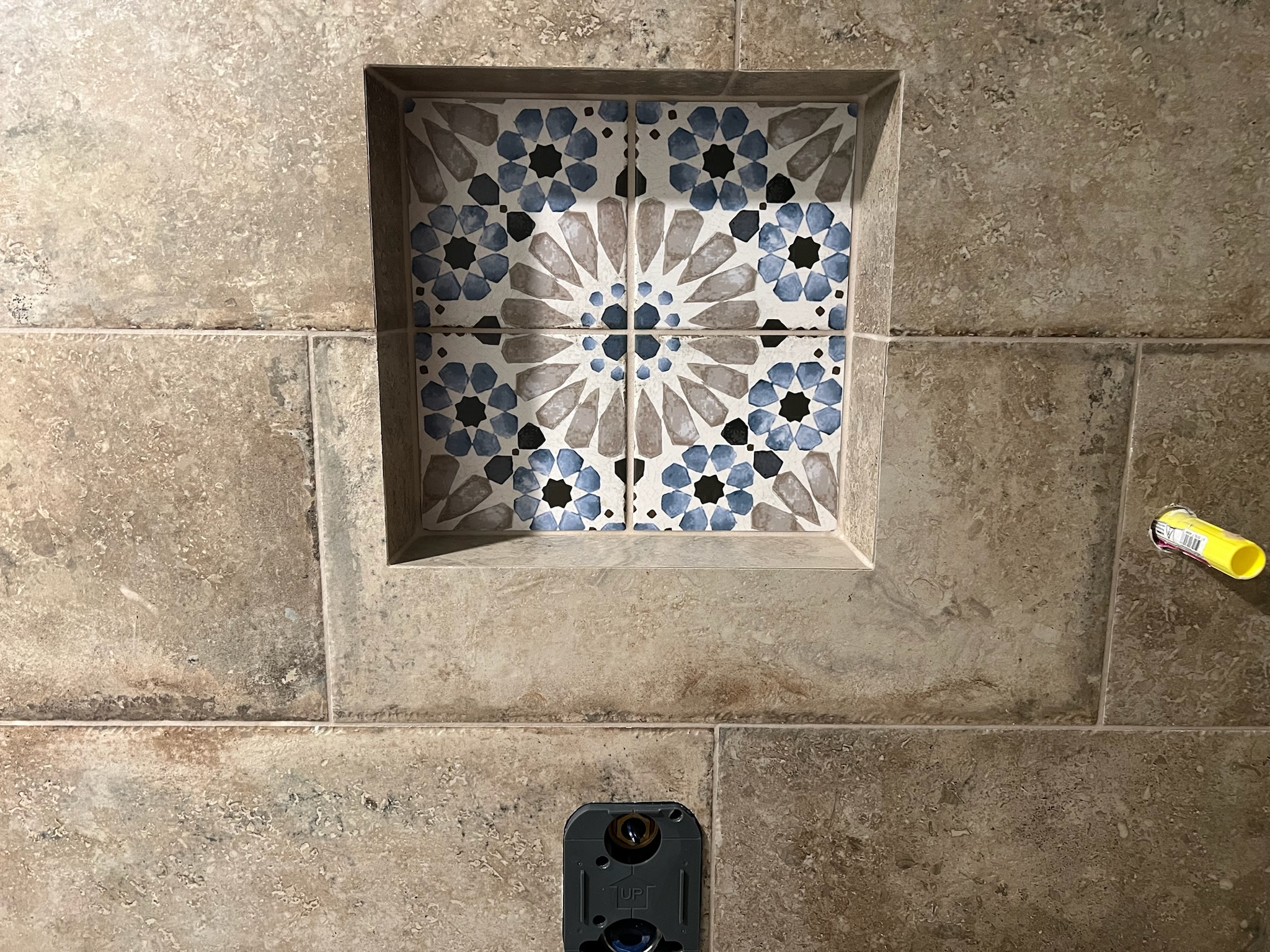 Close-up of beige bathroom wall tiles with a recessed square niche of blue and gray floral patterned tiles and a black fixture below.